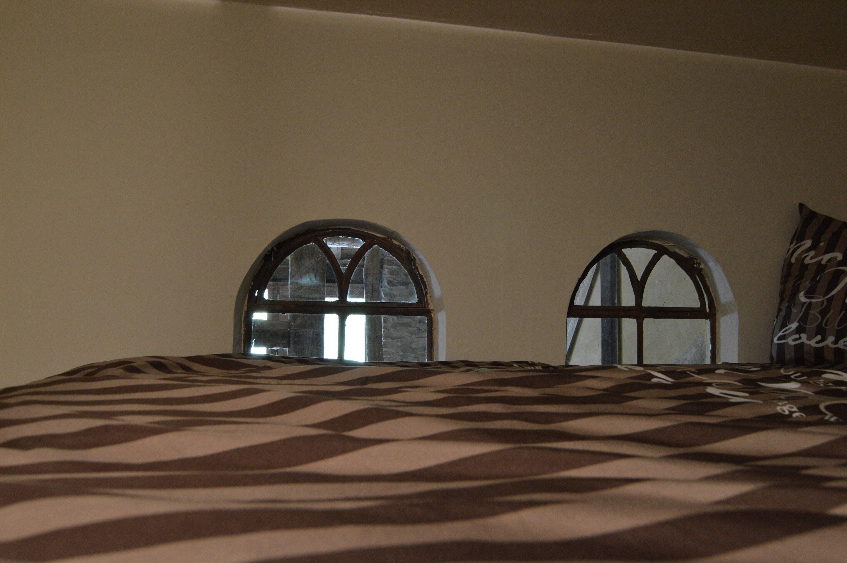 Ein Schlafzimmer mit einem gemusterten Bettbezug, zwei Rundbogenfenstern und einem Kopfkissen mit Schriftzug.