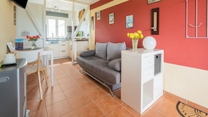 Un espace de vie compact comprenant un coin cuisine, un canapé gris, une petite table agrémentée d’un vase de fleurs et un téléviseur fixé au mur.