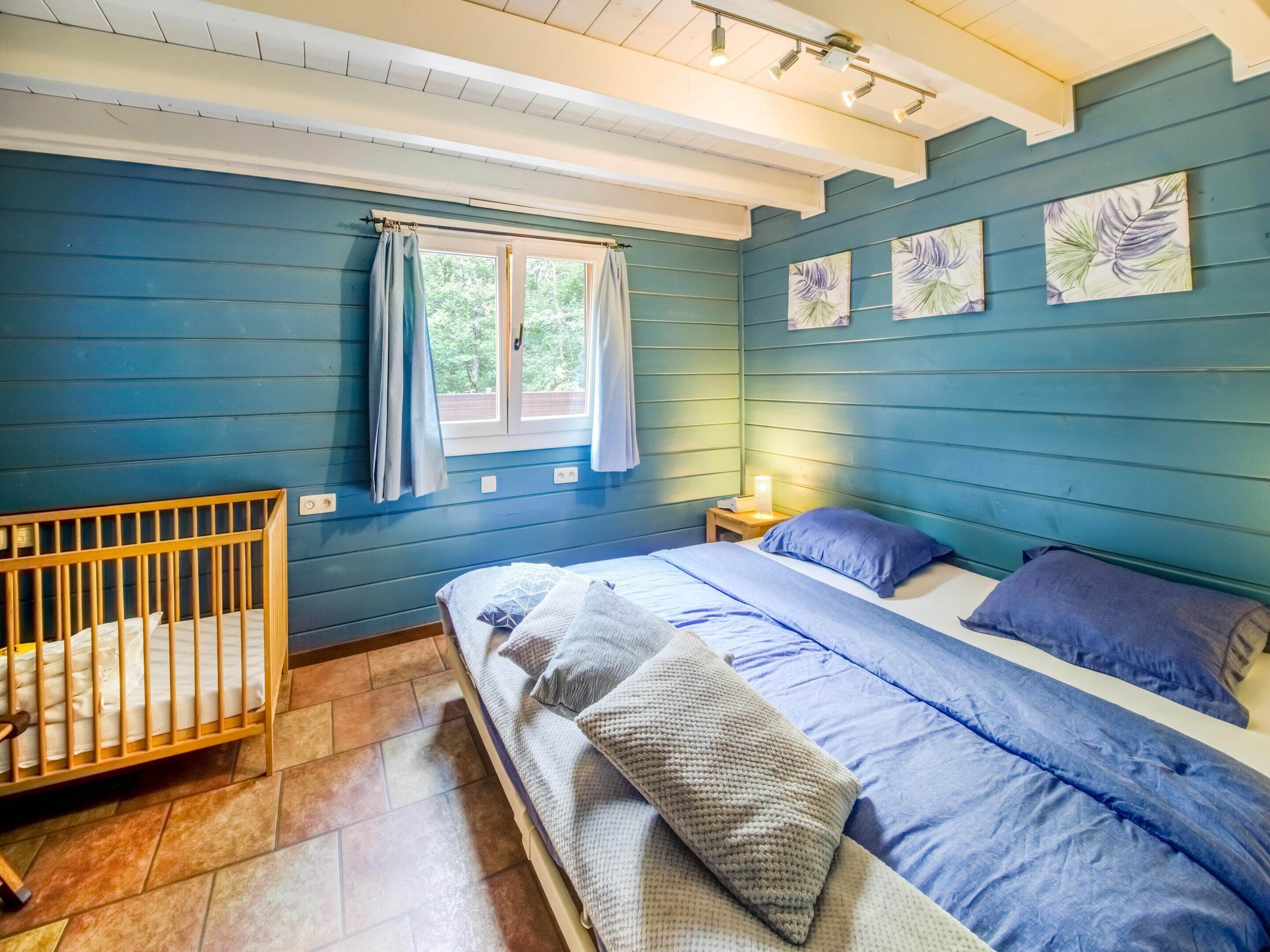 Un dormitorio con una cuna, una cama con ropa de cama azul, una mesita de noche y una ventana con cortinas.
