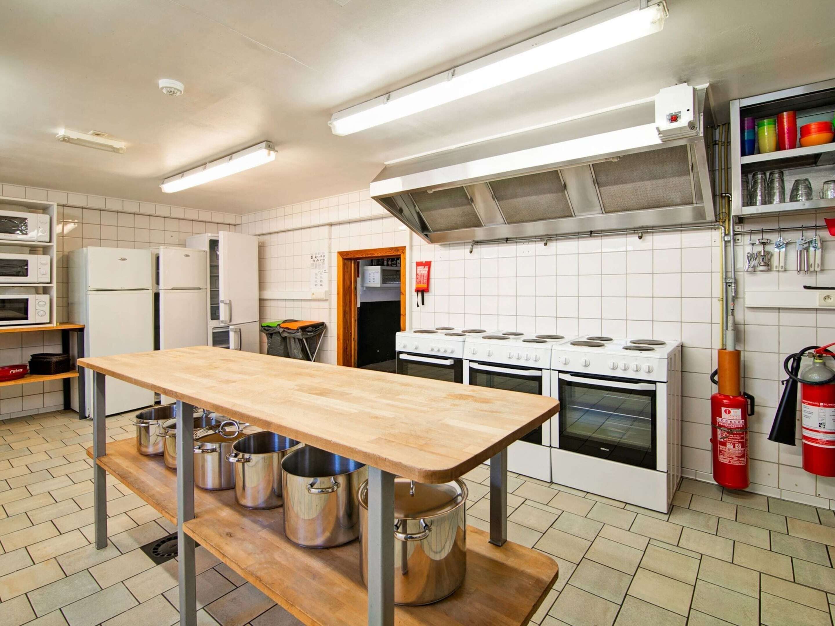 Una cocina con una mesa de madera, ollas de acero inoxidable, paredes de azulejos blancos, electrodomésticos blancos y un extintor.