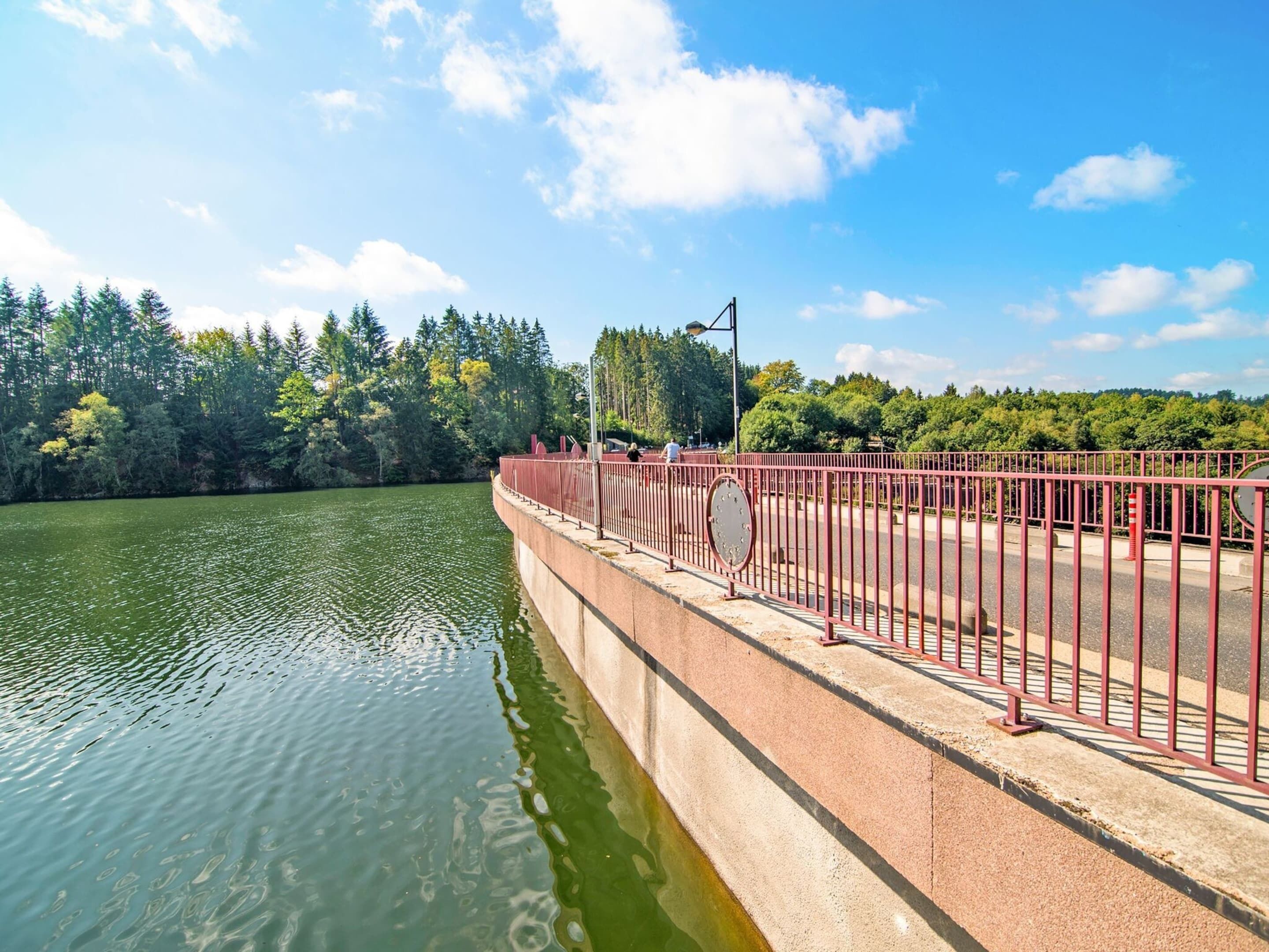 A concrete dam with a metal railing, a calm river, and a forested area in the background.
