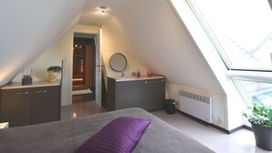 Un dormitorio moderno con techo inclinado, una cama con almohada morada, una mesita de noche, un espejo y una ventana con vistas al exterior.