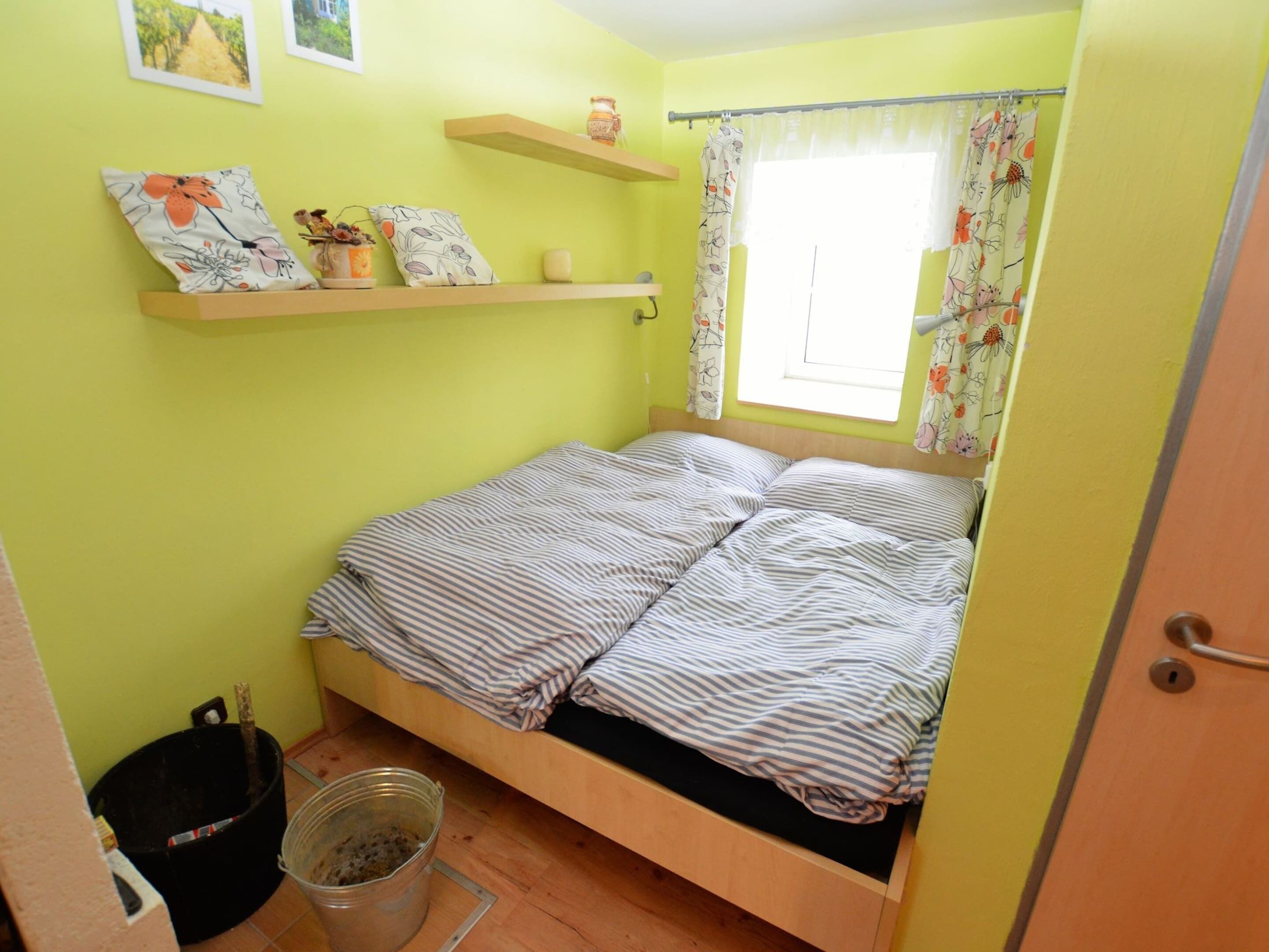A small, single-bed room with a wooden frame, a window with floral curtains, and a small shelf with decorative items.
