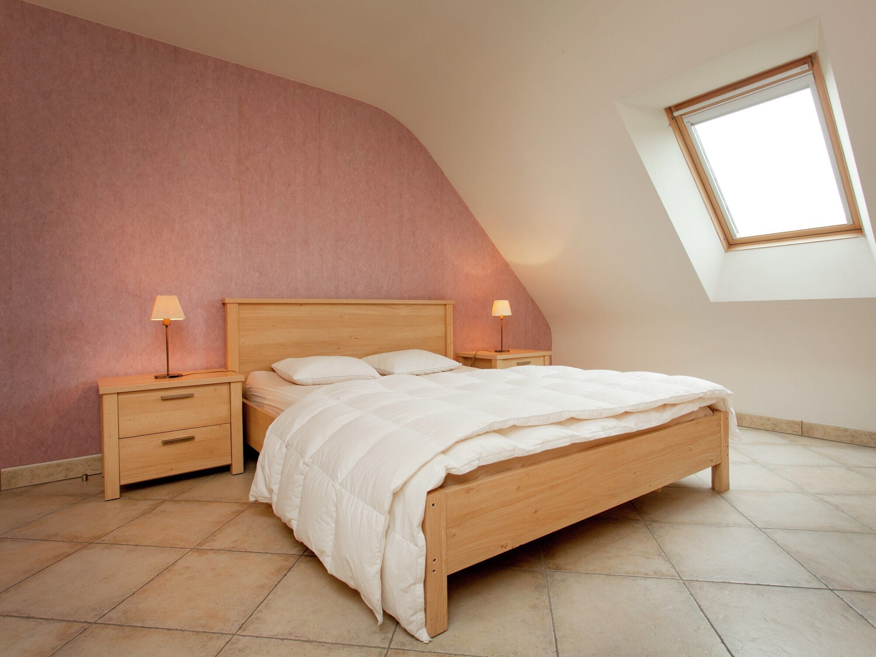 A bedroom with a sloped ceiling, a skylight, a wooden bed, two bedside tables with lamps, and a pink wall.