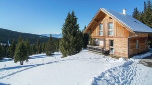 A wooden cabin with a balcony, surrounded by snow-covered ground and pine trees, with a clear blue sky. - Beautiful Holiday Home in Weinebene With Sauna (Frantschach-Sankt Gertraud)