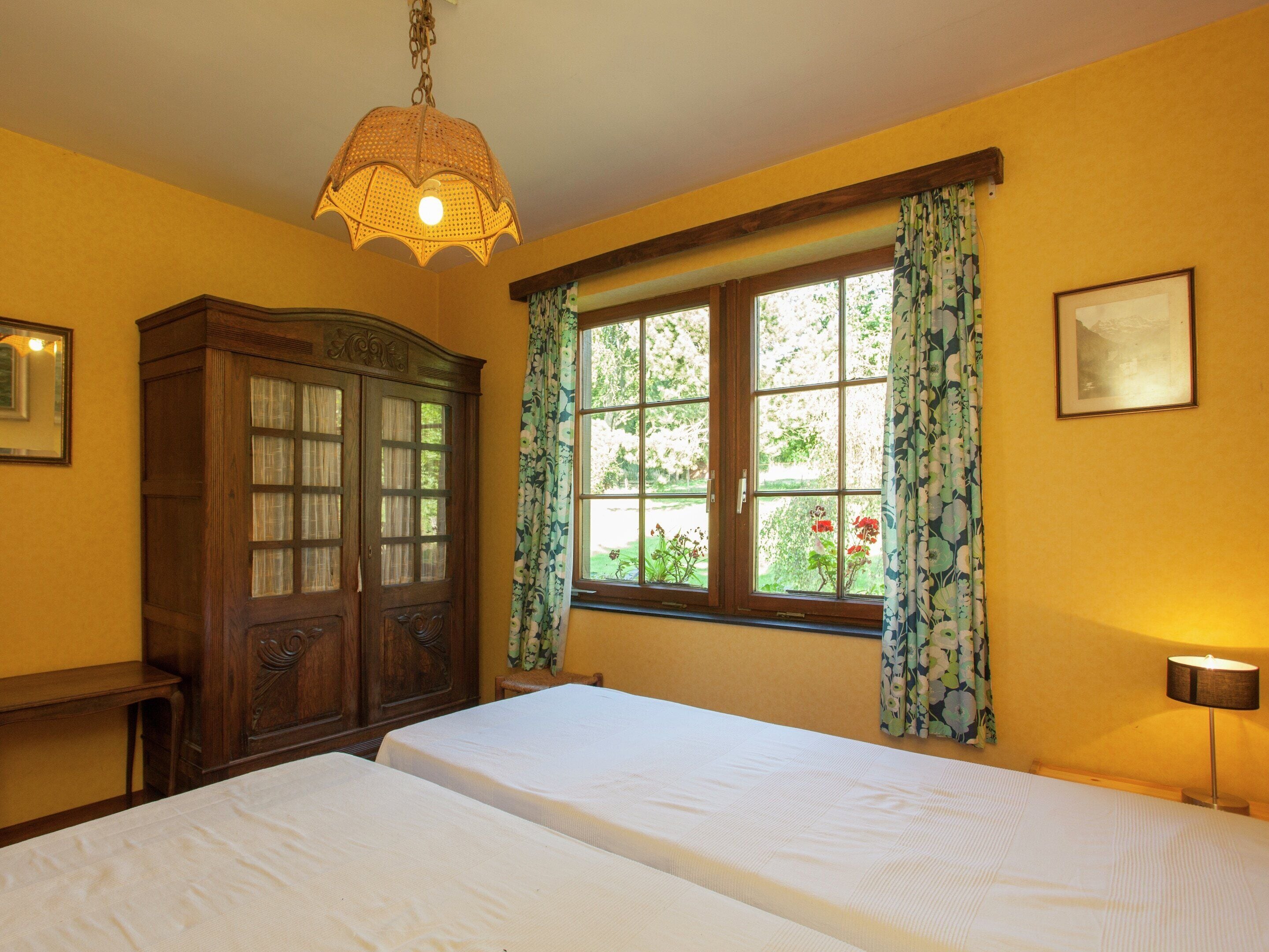 A room with two beds, a wooden cabinet, a window with floral curtains, and a hanging light fixture.