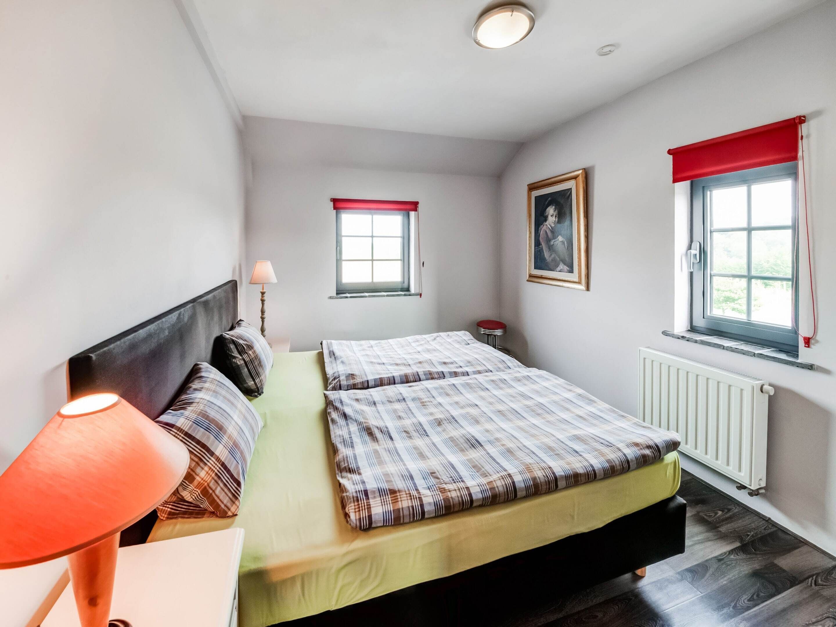 A bedroom with two beds, a nightstand, a lamp, a window with a red blind, a radiator, and a framed picture on the wall.