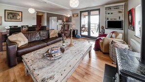 A living room with a brown leather sofa, a wooden coffee table with magazines, a patterned armchair, and a black fireplace. - Green Hillside Apartment-petit Wilogne (Houffalize)