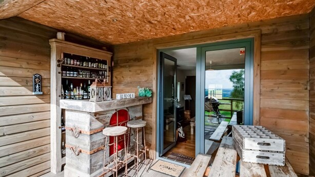A wooden interior with a bar area, stools, and a view through a glass door. - Green Hillside Apartment-petit Wilogne (Houffalize)