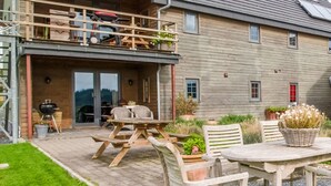 A two-story house with a patio area featuring a wooden table and chairs, a barbecue grill, and potted plants. - Green Hillside Apartment-petit Wilogne (Houffalize)