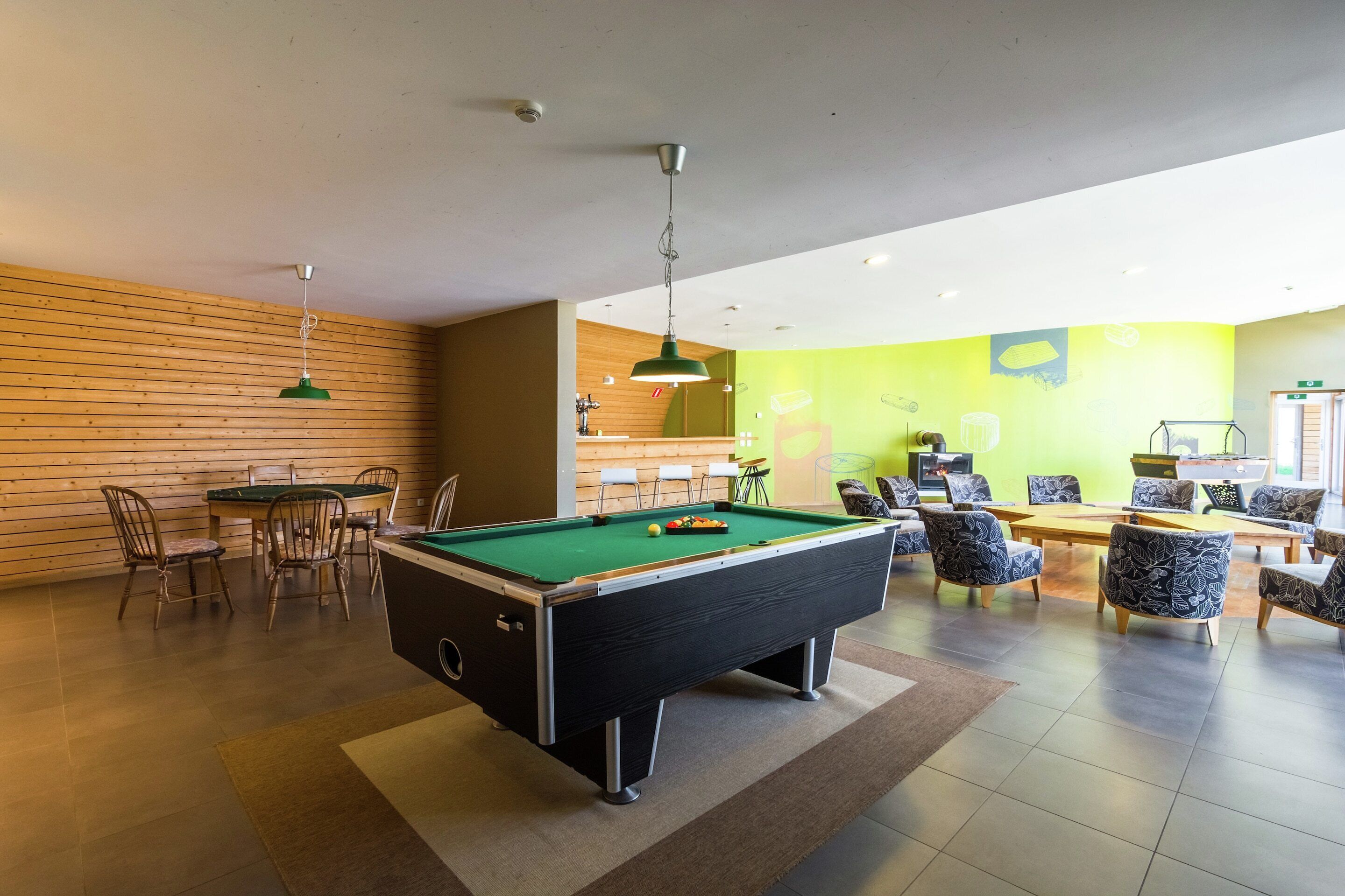 A pool table in a room with wooden paneling, a green wall, and a seating area with patterned chairs.
