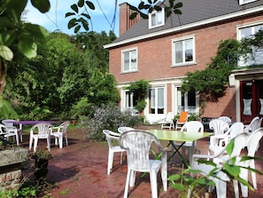 A patio area with white plastic chairs and tables, surrounded by greenery and a brick building.