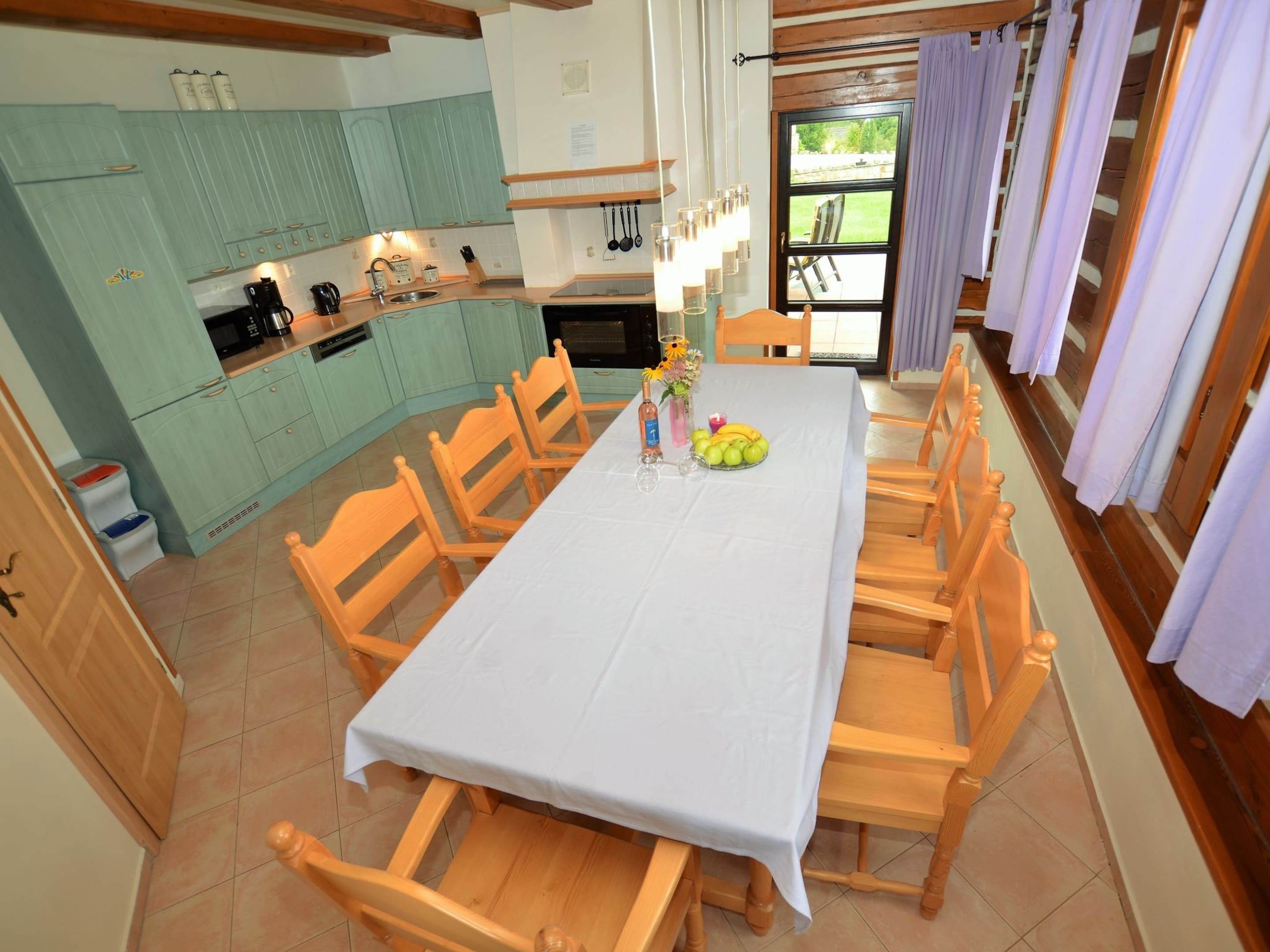 Un comedor con una mesa dispuesta para seis personas, sillas de madera, una cocina con armarios verde azulado y vistas al exterior a través de una ventana.
