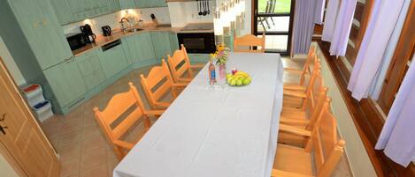 Un comedor con una mesa dispuesta para seis personas, sillas de madera, una cocina con armarios verde azulado y vistas al exterior a través de una ventana.