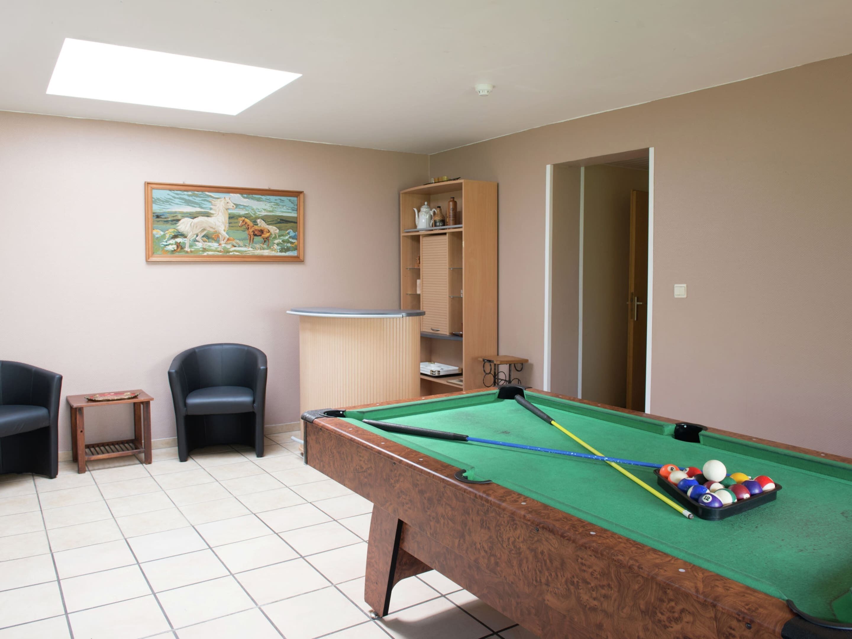 A pool table with balls, two chairs, a small table, a painting, and a shelf with items.