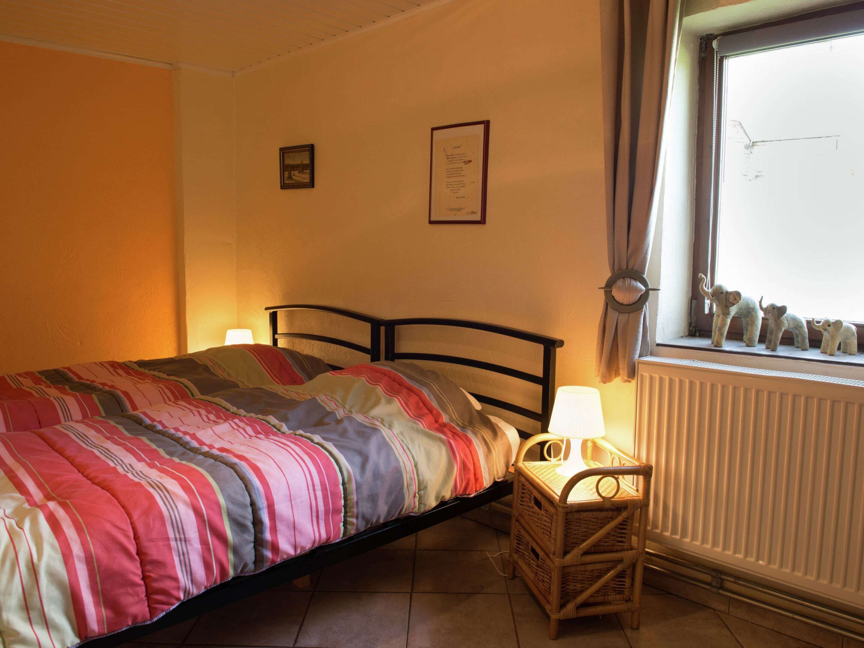 A bedroom with a bed, a nightstand, a lamp, a framed certificate on the wall, and a window with a view of the sky and a building.