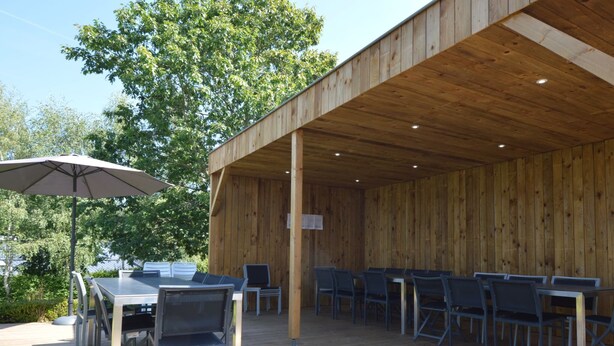 An outdoor wooden deck with tables and chairs, surrounded by trees. - Child Friendly Holiday Home in Waimes with Sauna & Hot Tub (Waimes)