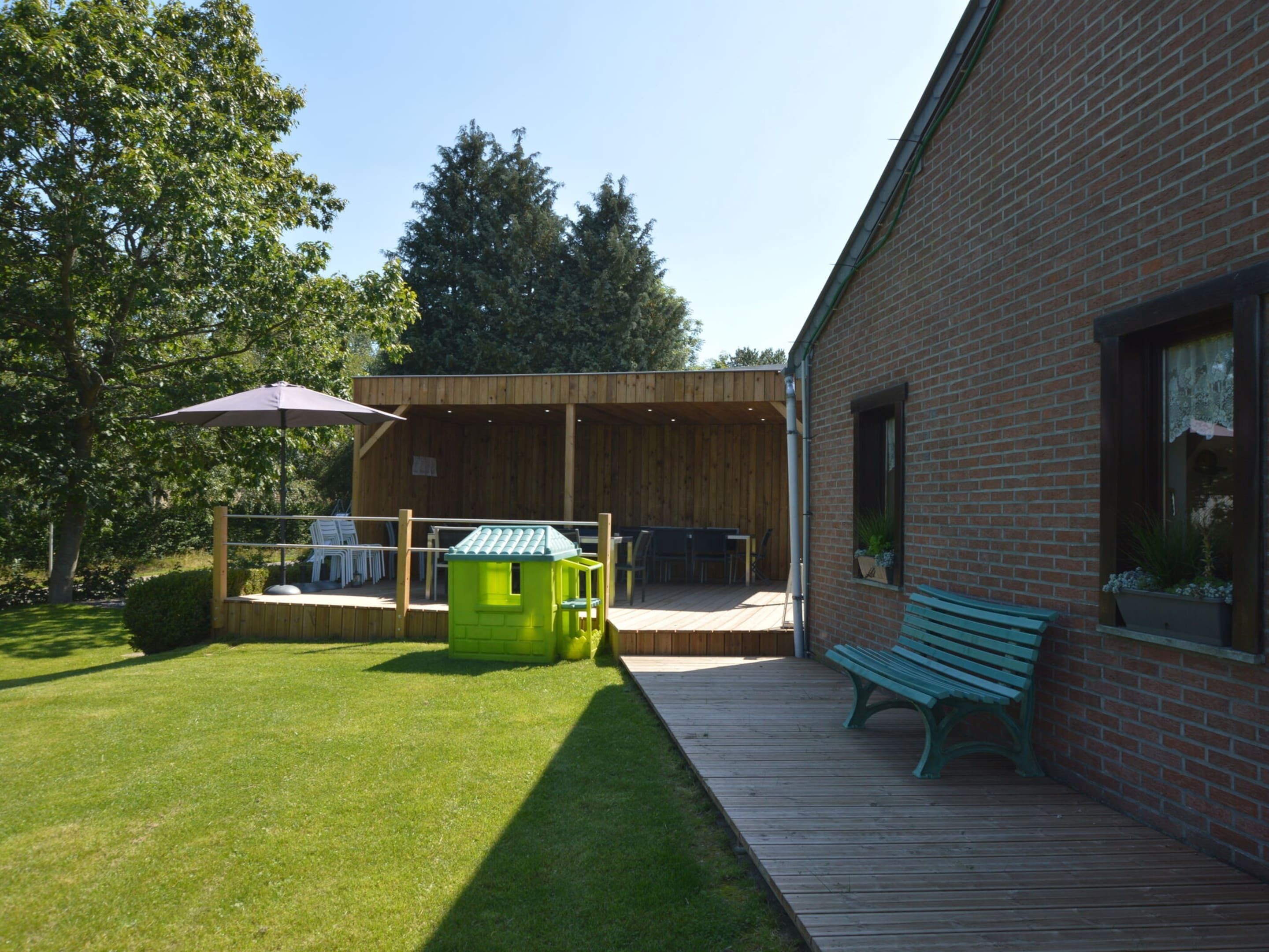 Eine Terrasse mit Holzboden, einem grünen Spielhäuschen, einer Bank und einem Backsteinhaus.
