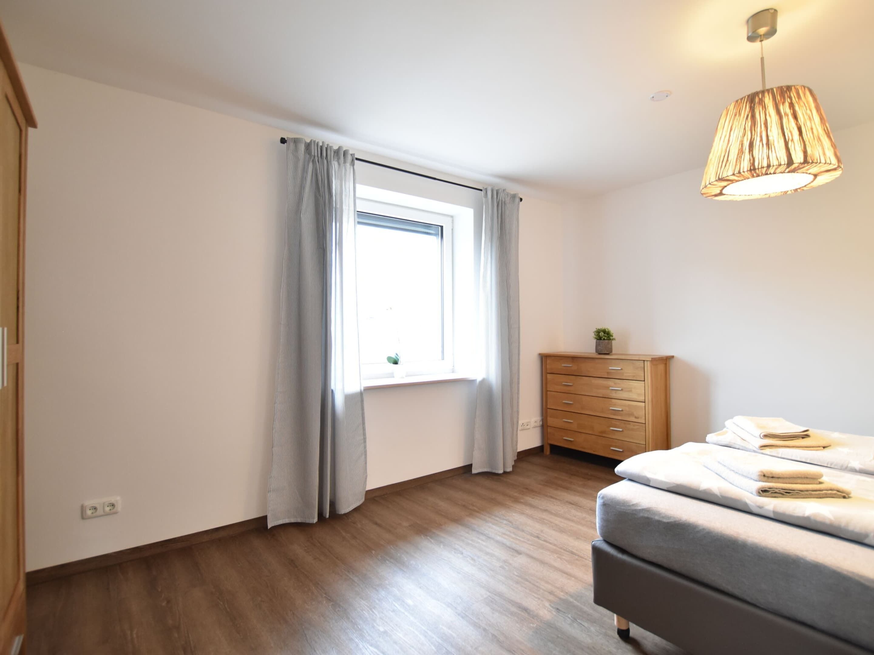 A bedroom with a wooden floor, a bed with a grey mattress, a wooden dresser, a window with curtains, and a hanging lamp.