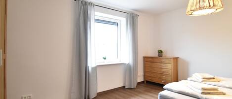 Un dormitorio con piso de madera, una cama con colchón gris, un aparador de madera, una ventana con cortinas y una lámpara colgante.