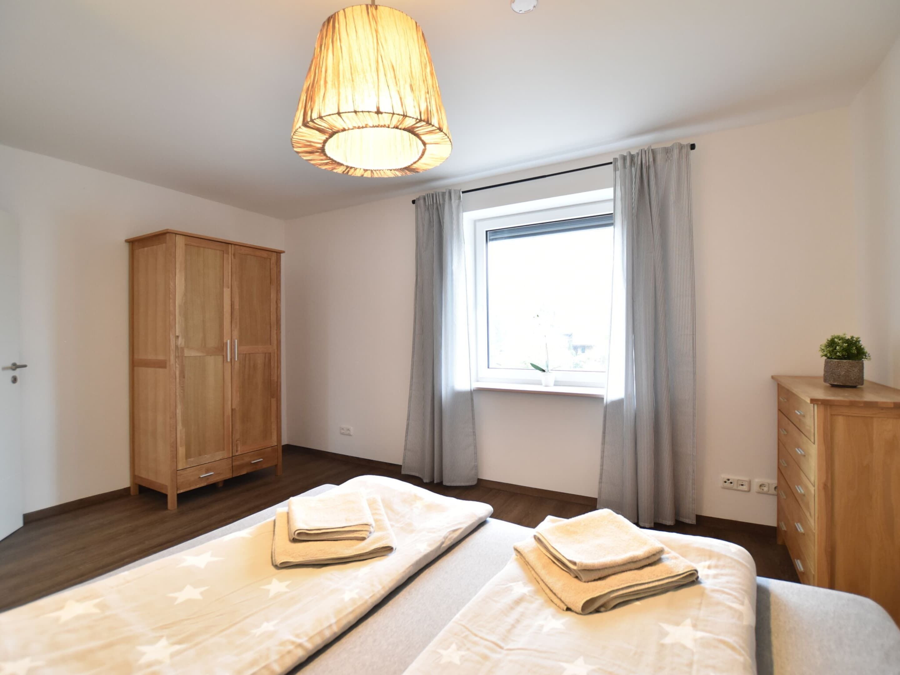 A bedroom with two beds, a wooden wardrobe, a window with curtains, and a hanging light fixture.
