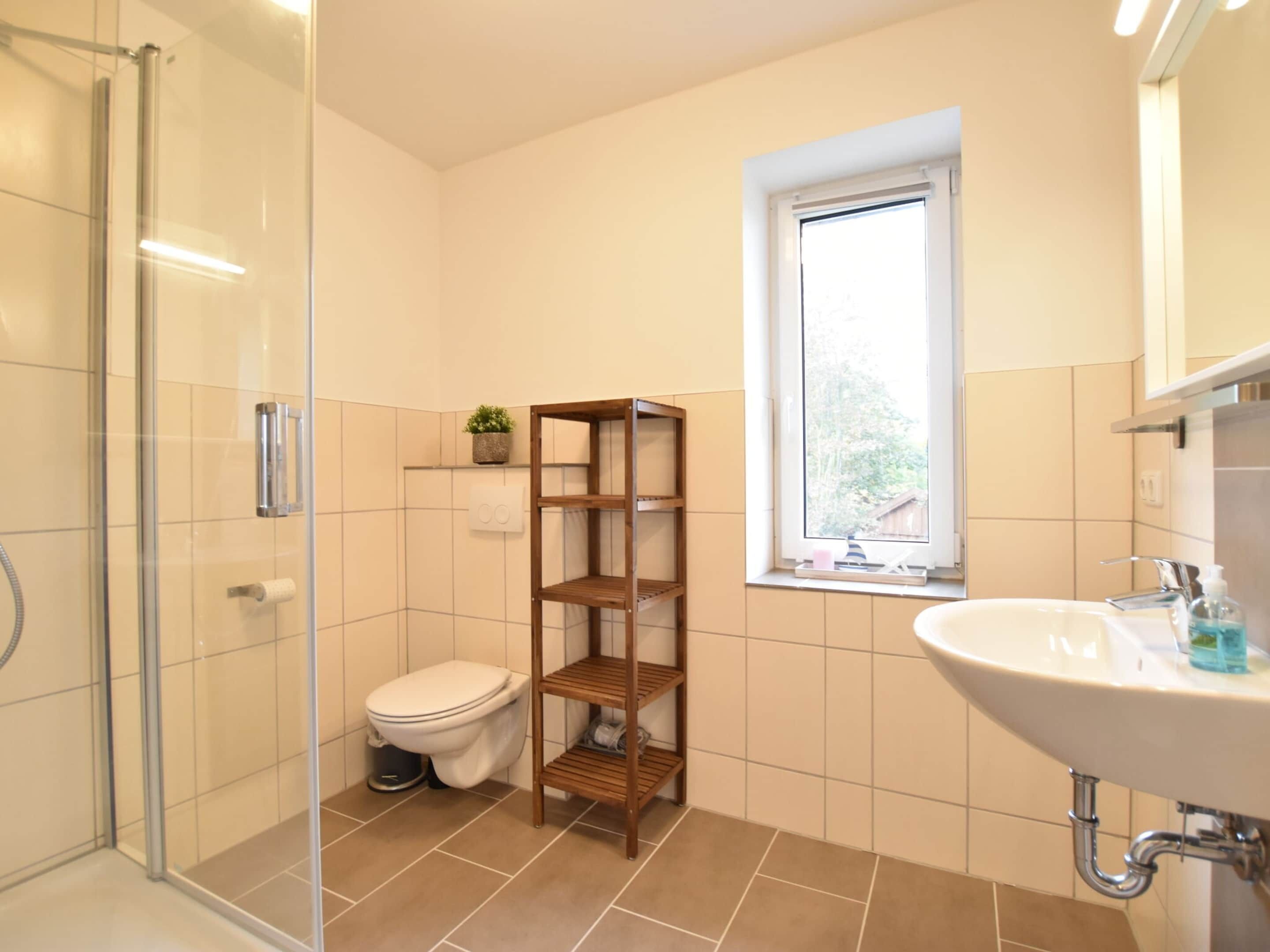 Ein Badezimmer mit Duschkabine aus Glas, Toilette, Holzregal, Fenster und Waschbecken mit Spiegel.