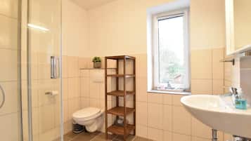 Ein Badezimmer mit Duschkabine aus Glas, Toilette, Holzregal, Fenster und Waschbecken mit Spiegel.
