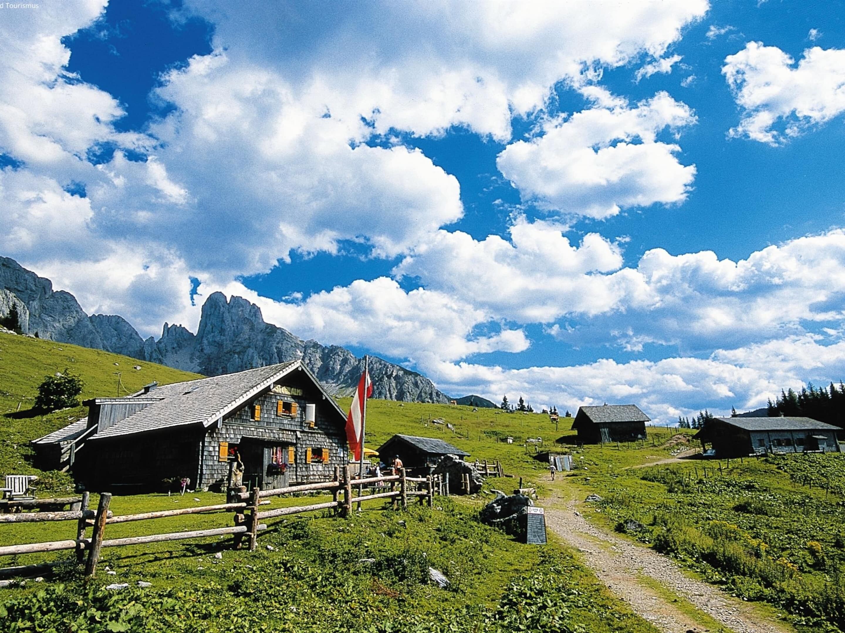 A traditional wooden chalet with a sloped roof, surrounded by a wooden fence, set against a backdrop of mountains and a clear sky.