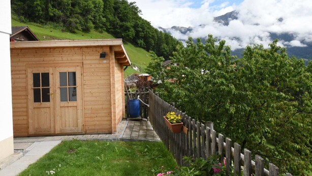 Une cabane en bois avec une porte et des fenêtres en bois, un petit porche et une vue sur une colline verdoyante et des montagnes en arrière-plan.