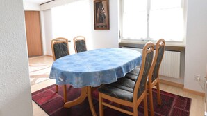 Un coin repas avec une table ronde recouverte d’une nappe bleue à motifs floraux, quatre chaises en bois avec des assises rembourrées, un tapis rouge à motifs et une fenêtre donnant sur l’extérieur.