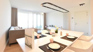 A modern living room with a sofa, dining table set with plates, cups, and cutlery, and a large window with curtains. - Bright Coastal Family Escape (De Panne)