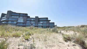 A modern multi-story building with a flat roof and large windows, situated on a sandy dune with sparse vegetation. - Bright Coastal Family Escape (De Panne)