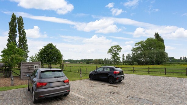 Two cars parked on a cobblestone driveway in a rural setting with green fields and trees in the background.