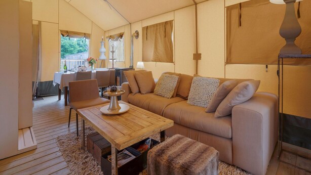 A cozy living room with a sofa, coffee table, and a fireplace. There is a dining area with a table and chairs, and a view of the outdoors through large windows. - Cozy Tent Lodge in Pittem With Veranda,barbecue (Pittem)