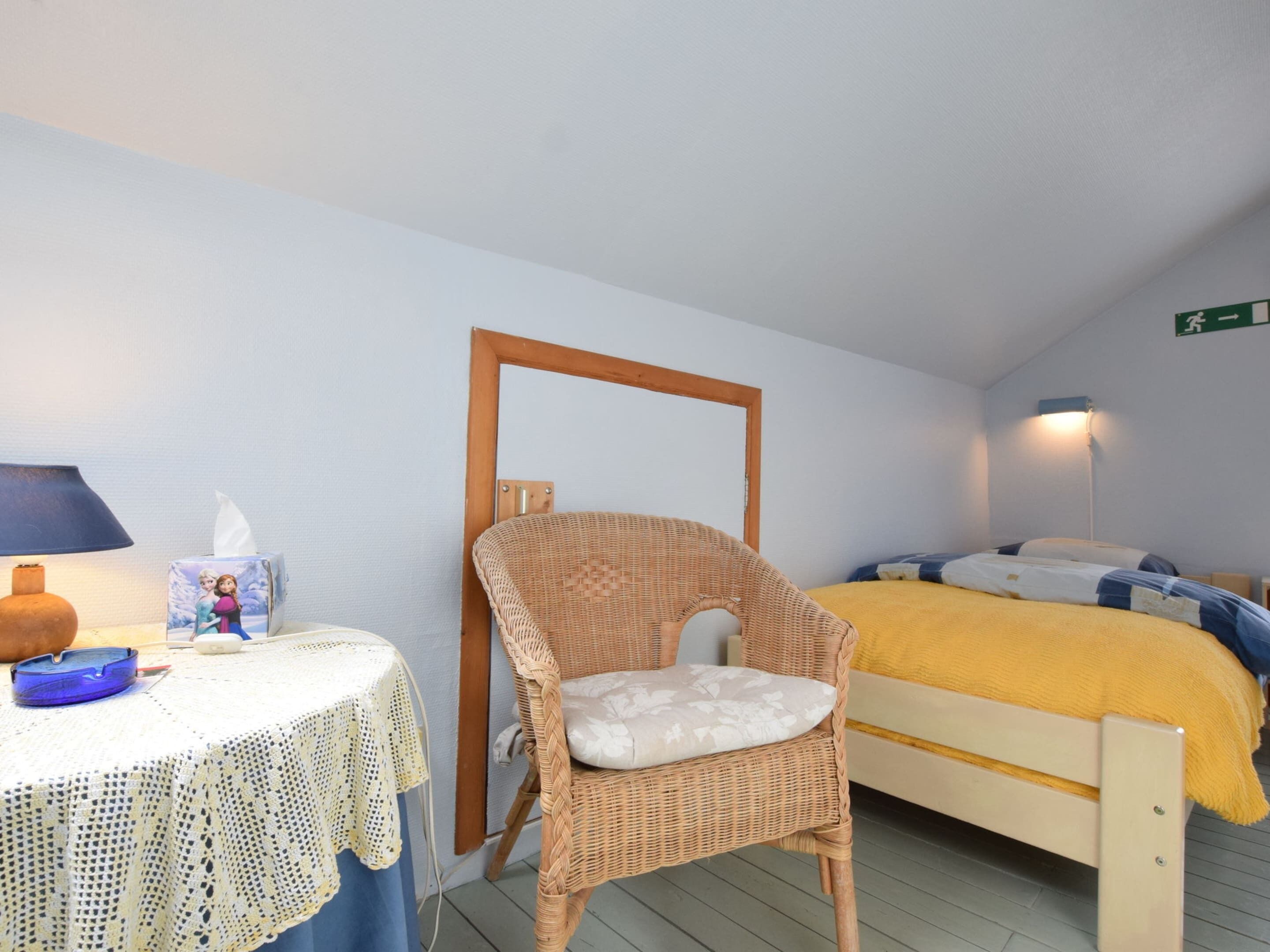 Ein kleiner, gemütlicher Raum mit einem Korbstuhl, einem Bett mit gelber Decke, einem Tisch mit weißer Spitzenleinentischdecke und einer Lampe.