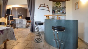 A cozy living room with a fireplace, a bar area with stools, and a dining table with a floral tablecloth. - Lovely Spot With Plenty of Outdoor Space (Sainte-Ode)