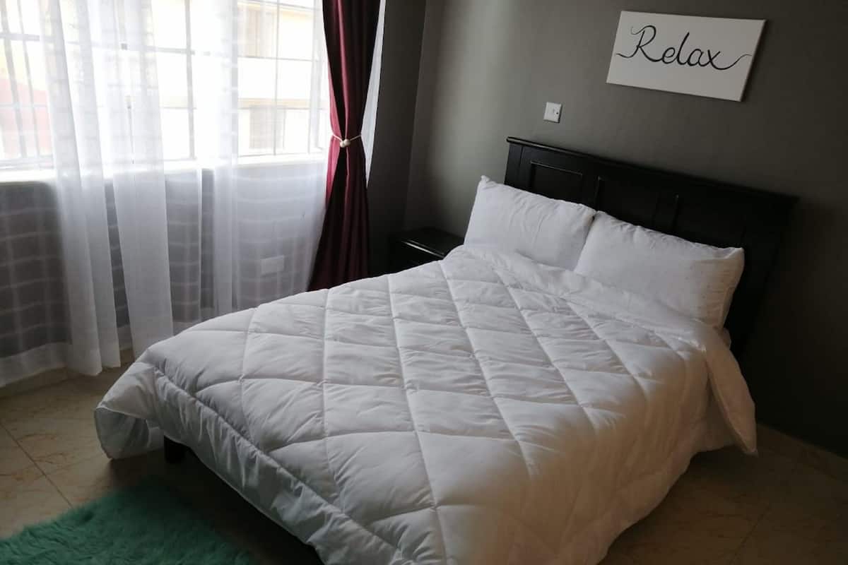 A neatly made bed with a white quilted bedspread and pillows, a green rug, and a 'Relax' sign on the wall.