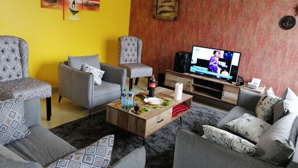 A living room with a sofa set, a coffee table with items, a television, and artwork on the walls. - Yurana Resort Kenya (Kitengela)