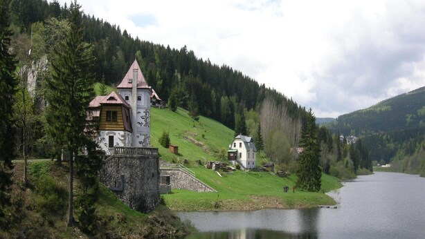 A castle-like building with a stone base and a red roof, situated by a calm lake surrounded by forested hills.