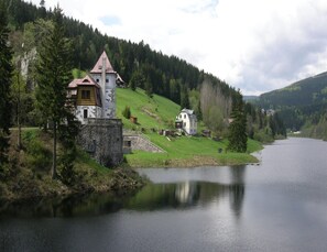 Ein schlösserähnliches Gebäude mit steinernem Fundament und rotem Dach, gelegen an einem stillen See, umgeben von bewaldeten Hügeln.