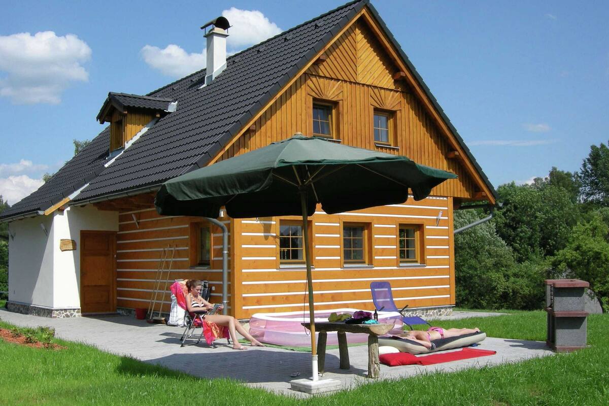 Una cabaña de troncos con techo inclinado, una chimenea y un porche con un paraguas verde. Hay dos sillones de descanso y una pequeña mesa con objetos encima.