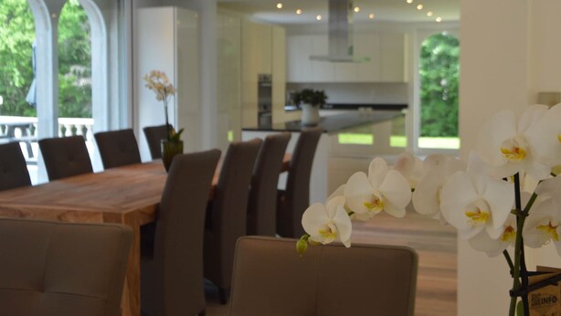 Een moderne eethoek met een houten tafel en stoelen, een keuken op de achtergrond en witte orchideeën op de tafel.