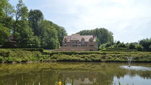 Een groot, historisch gebouw met een symmetrische gevel, gelegen naast een rustig vijver met een fontein.