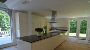 Een moderne keuken met een groot eiland, witte kasten en uitzicht op de omgeving door grote ramen.