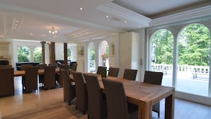 Ruime eetruimte met een grote houten tafel en meerdere stoelen, grote ramen met rondbogen en uitzicht op groen.