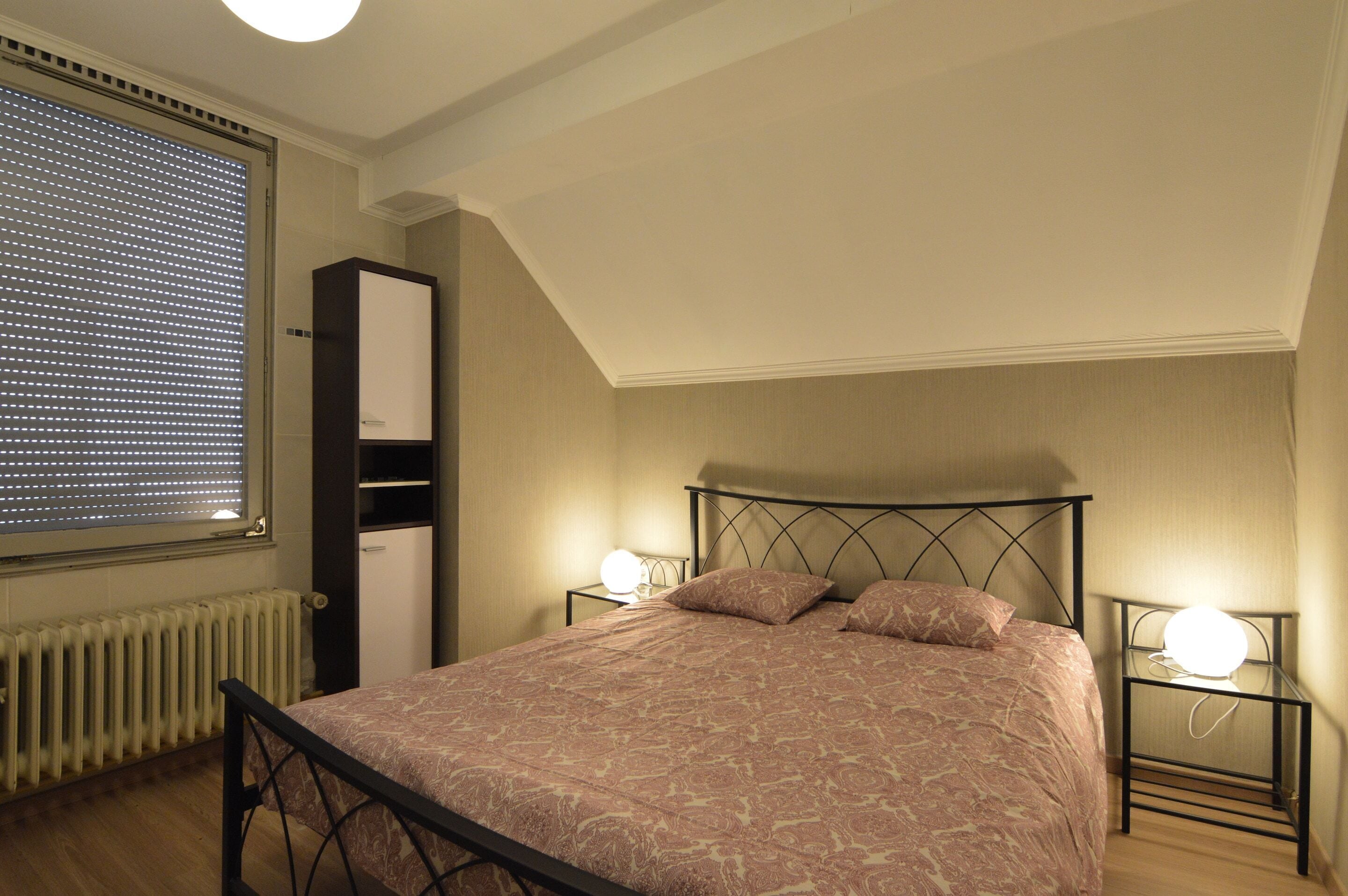 A bedroom with a bed, a radiator, a window with blinds, a wardrobe, and bedside lamps.