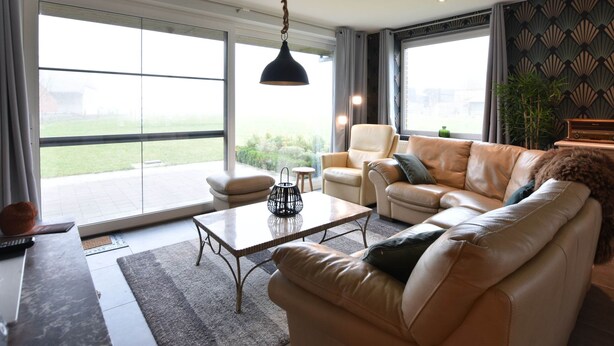 A spacious living room with a large window, a leather sofa, a coffee table, and a hanging light fixture. - Holiday Home in Alveringem Near Beach (Alveringem)