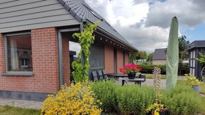 A brick house with a covered patio, a tall green plant, and yellow flowers in the garden. - Holiday Home in Alveringem Near Beach (Alveringem)