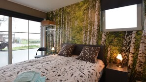 A bedroom with a large bed, two bedside lamps, a window with a view, and a wall with a nature-themed wallpaper. - Holiday Home in Alveringem Near Beach (Alveringem)