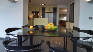 A modern dining area with a glass table and black chairs, a fruit bowl on the table, and a kitchen in the background. - Beautiful Villa for Groups With Swimming Pool, Sauna and Wifi (Zottegem)